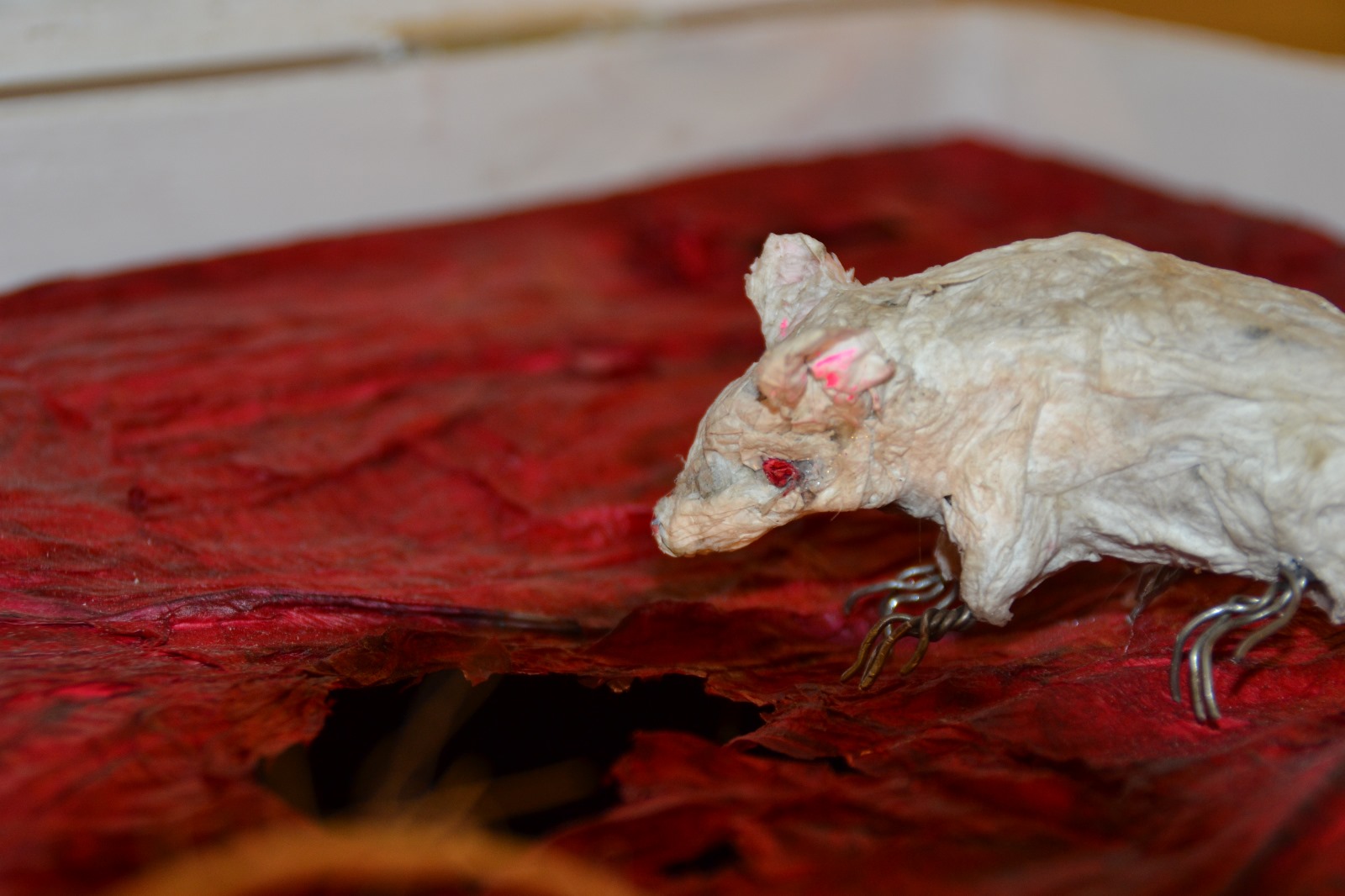 Close-up of a papier-mâché and wire sculpture of a mouse peeking through a crack in a red base.